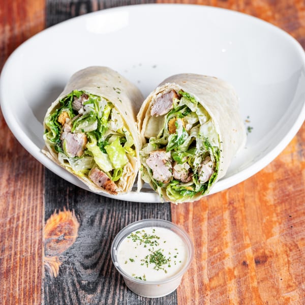 Two halves of a chicken Caesar wrap with dressing, served on a white plate with a side of sauce.